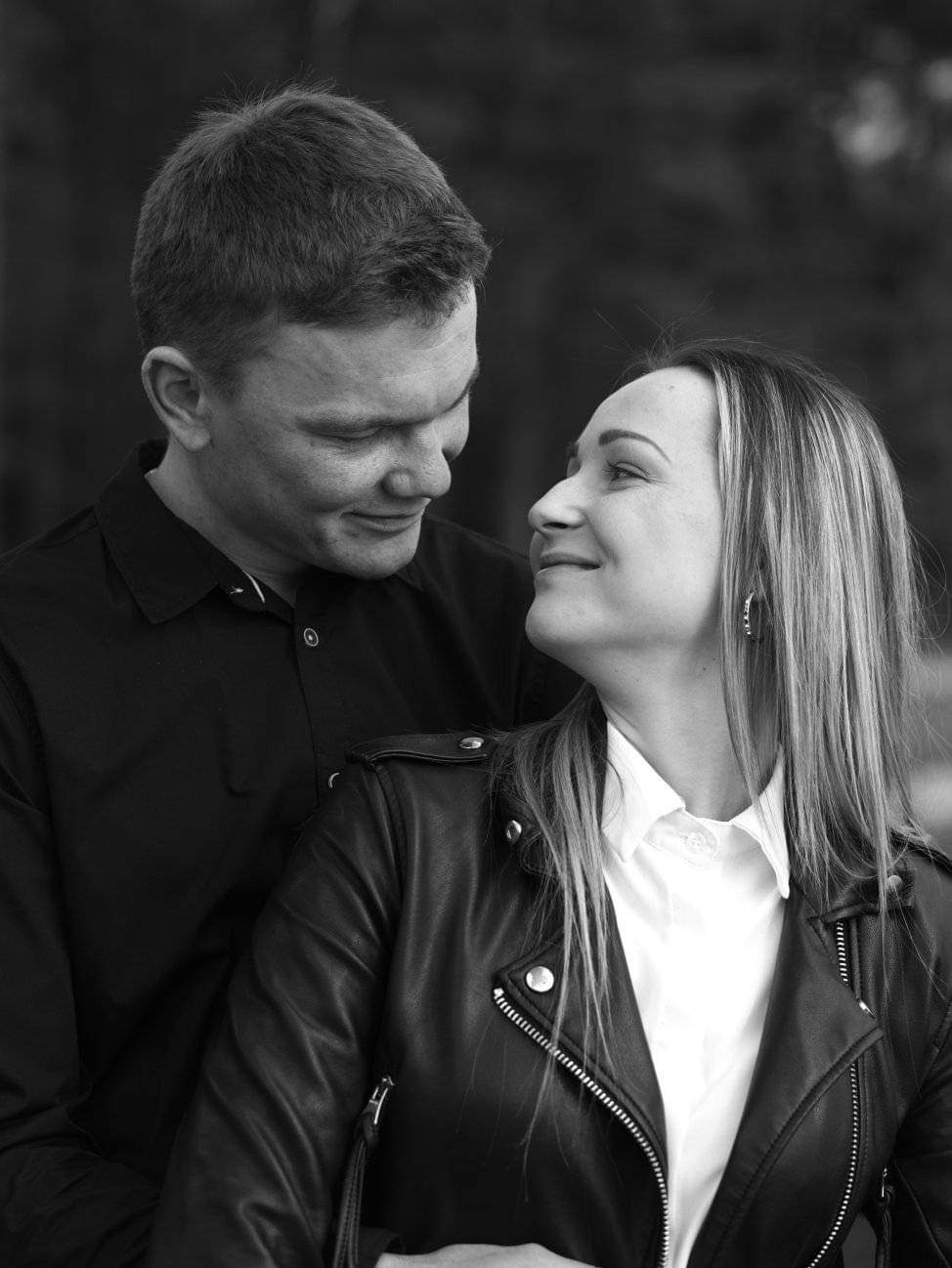 Czarno-białe zdjęcie pary: mężczyzna spogląda z uśmiechem na kobietę w skórzanej kurtce, która patrzy na niego z czułością.