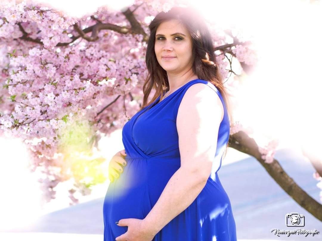 Kobieta w niebieskiej sukience ciążowej pozuje na tle kwitnącej wiśni w promieniach słońca, dłoń na brzuchu.