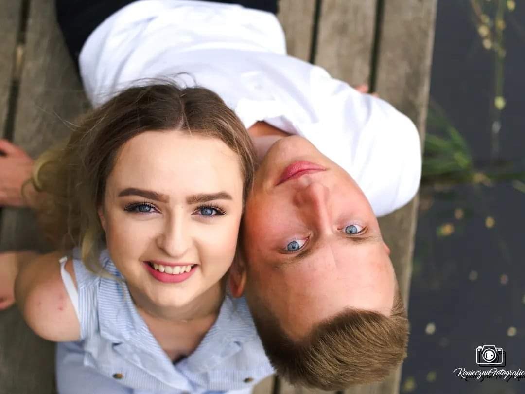 Młoda uśmiechnięta para leżąca na drewnianym pomoście, widok z góry, w tle woda.