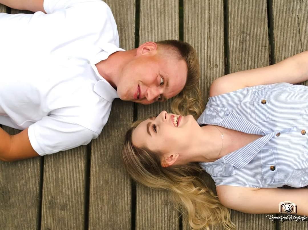 Uśmiechnięta para leżąca na drewnianym pomoście, on w białej koszulce polo, ona w bluzce w niebieskie paski, patrzą na siebie z góry.