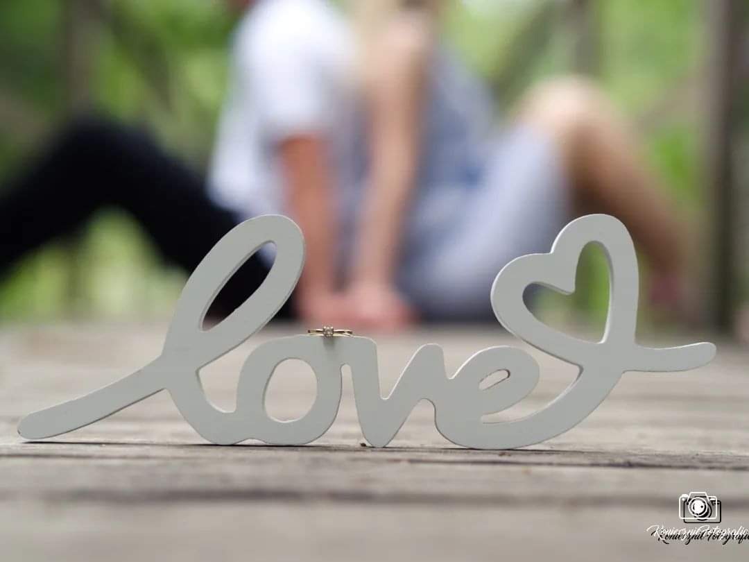 Drewniany napis LOVE z pierścionkiem zaręczynowym na literze 'V', w tle para siedząca na drewnianym pomoście, rozmyte tło zieleni.