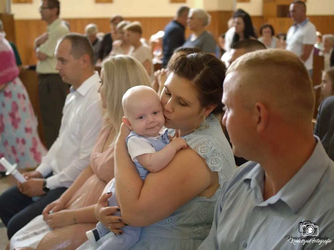Matka całuje w policzek ubranego na niebiesko niemowlaka na tle siedzących osób w kościele, prawdopodobnie podczas uroczystości religijnej.