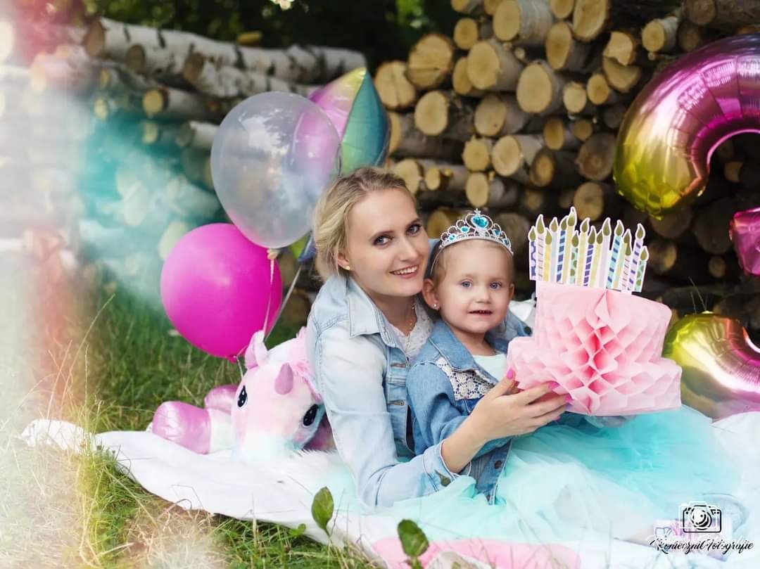 Uśmiechnięta mama obejmuje córeczkę w koronie, siedzącą na kocu z tortem-dekoracją i balonami na tle stosu drewna.