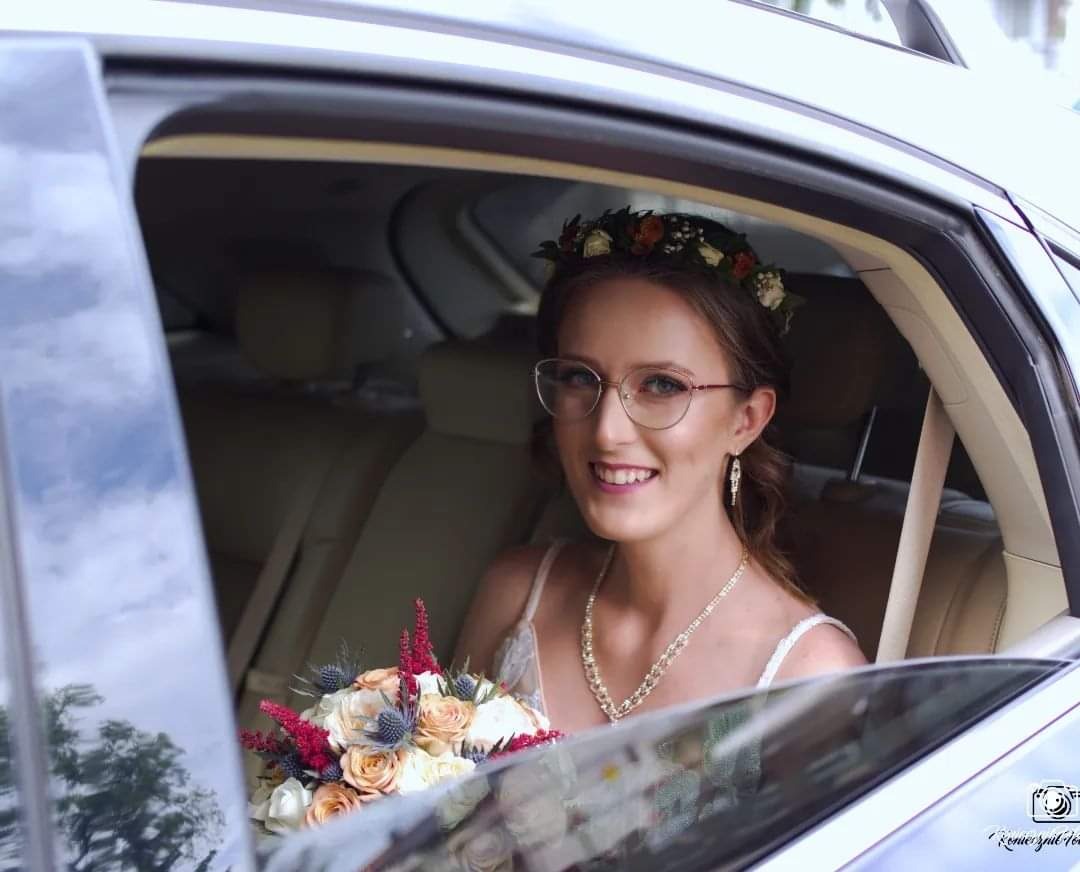 Uśmiechnięta panna młoda w okularach, z bukietem i wiankiem na głowie, siedzi w samochodzie, w tle widoczne beżowe skórzane siedzenia.