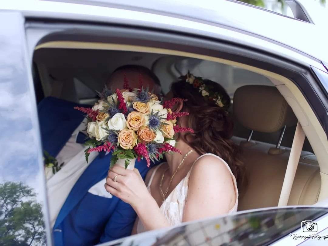 Para młoda w samochodzie, pocałunek zasłonięty bukietem ślubnym z róż, ostu i różowych kwiatów, panna młoda ma wianek na włosach.