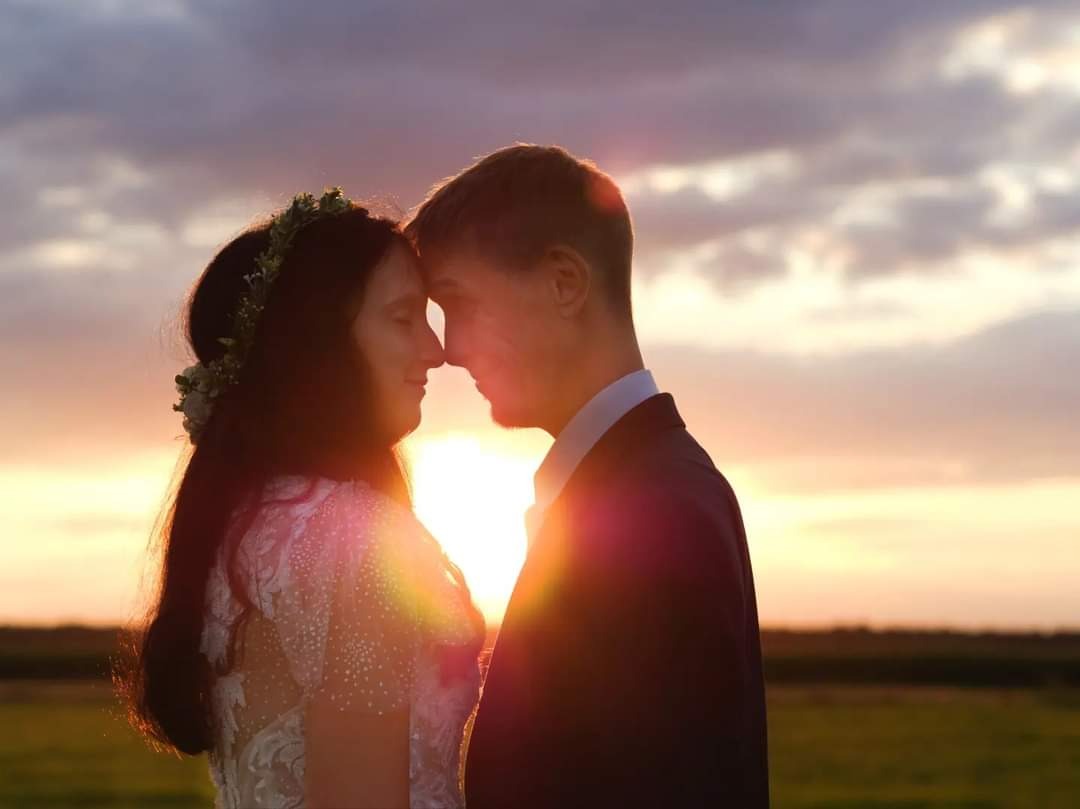 Para młoda z Grójca, stykająca się czołami na tle zachodzącego słońca, delikatne światło podkreśla koronę panny młodej.