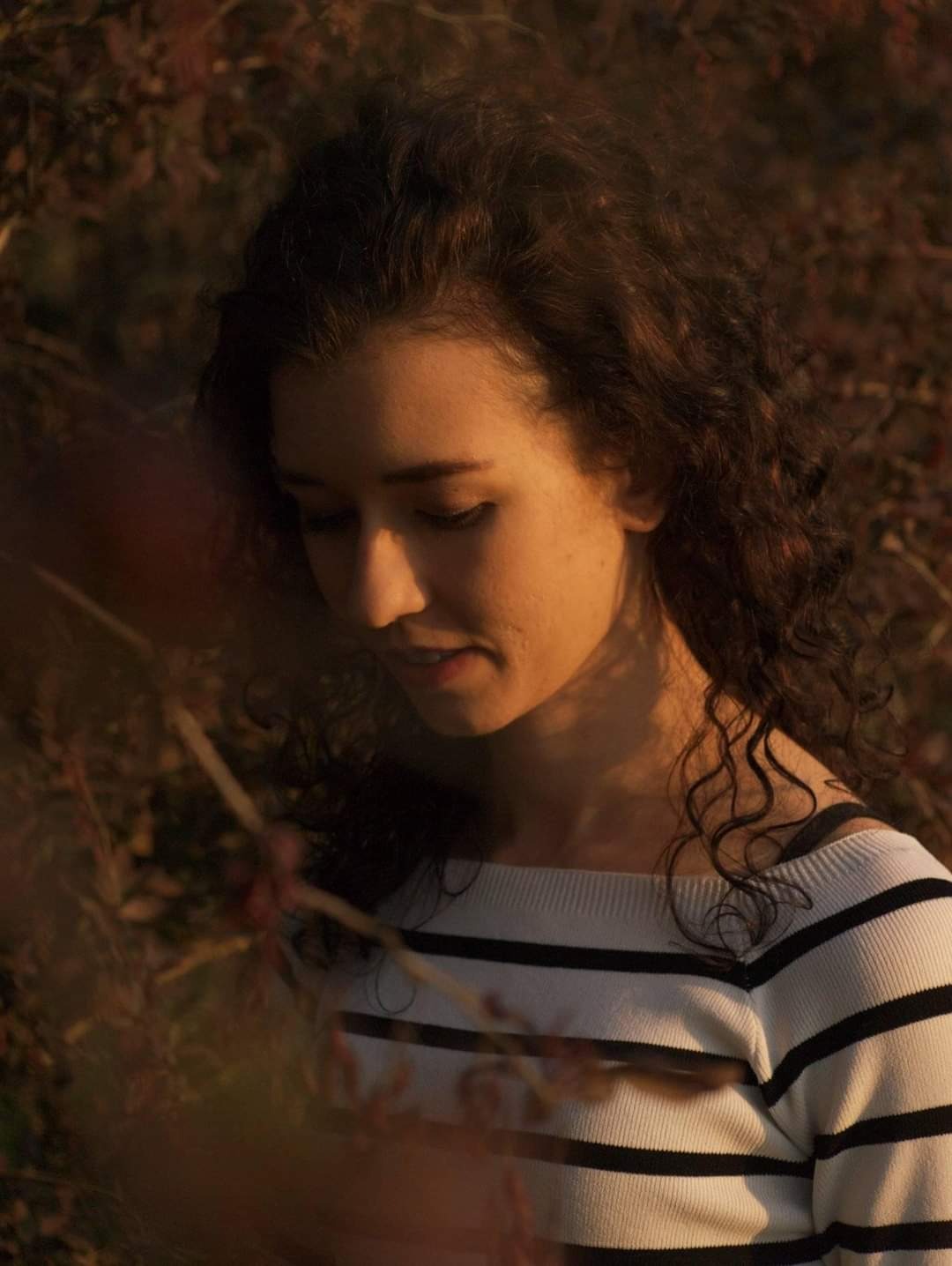 Portret młodej kobiety z kręconymi włosami, spoglądającej w dół, w swetrze w czarno-białe paski, na tle jesiennej roślinności w ciepłym świetle.