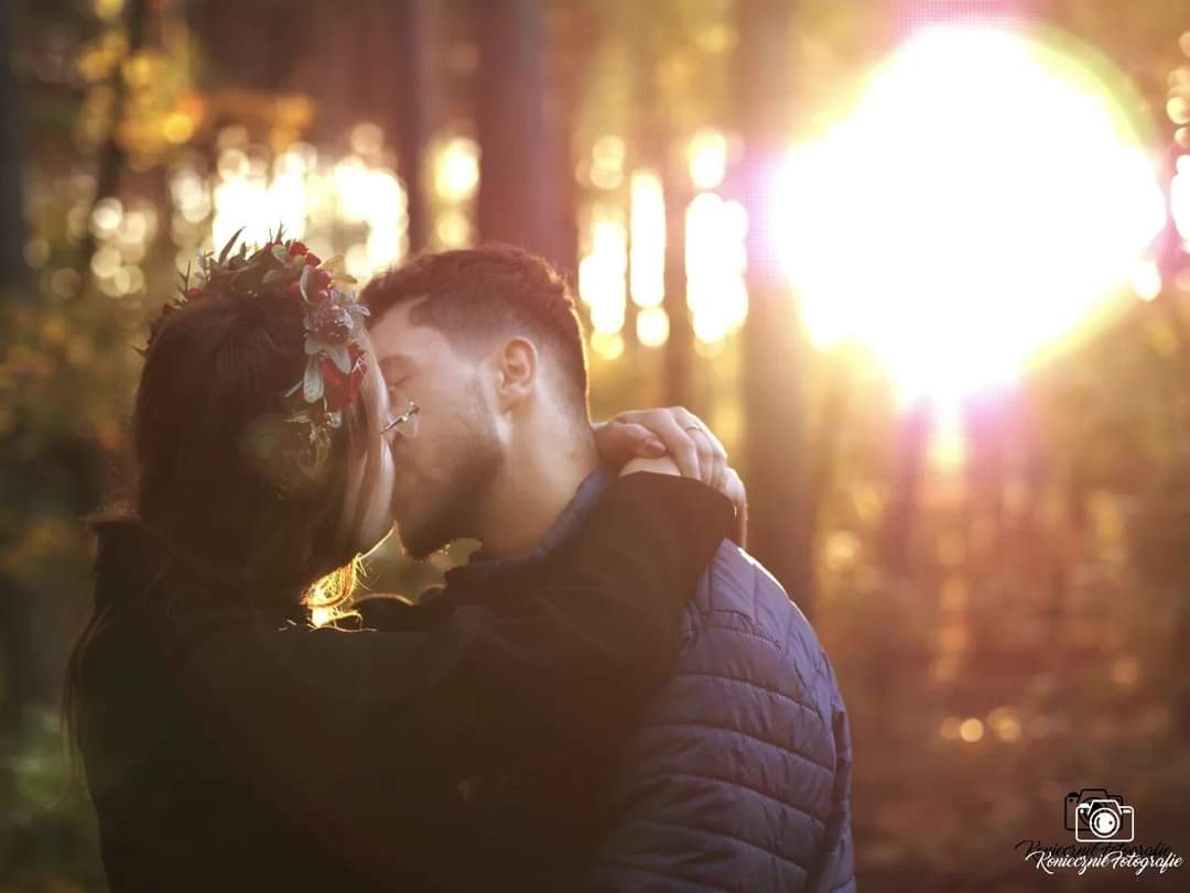 Zakochana para całuje się w lesie oświetlonym zachodzącym słońcem, kobieta ma wianek z kwiatów na głowie, widoczne logo fotografa w prawym dolnym rogu.