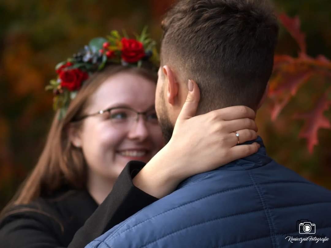 Uśmiechnięta kobieta w wianku z czerwonych róż obejmuje mężczyznę, na jej palcu widoczny pierścionek zaręczynowy, jesienne tło.