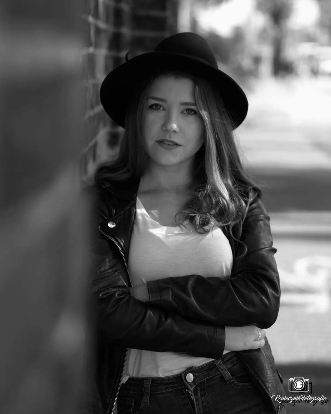 Portret młodej kobiety w czarnym kapeluszu i skórzanej kurtce, oparta o ceglaną ścianę, czarno-białe zdjęcie z rozmytym tłem miejskim.