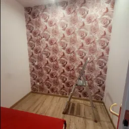 Pokój z tapetą w różowe róże w trakcie remontu, widoczna drabina aluminiowa, gniazdka elektryczne i wystające kable, część czerwonego łóżka w rogu.