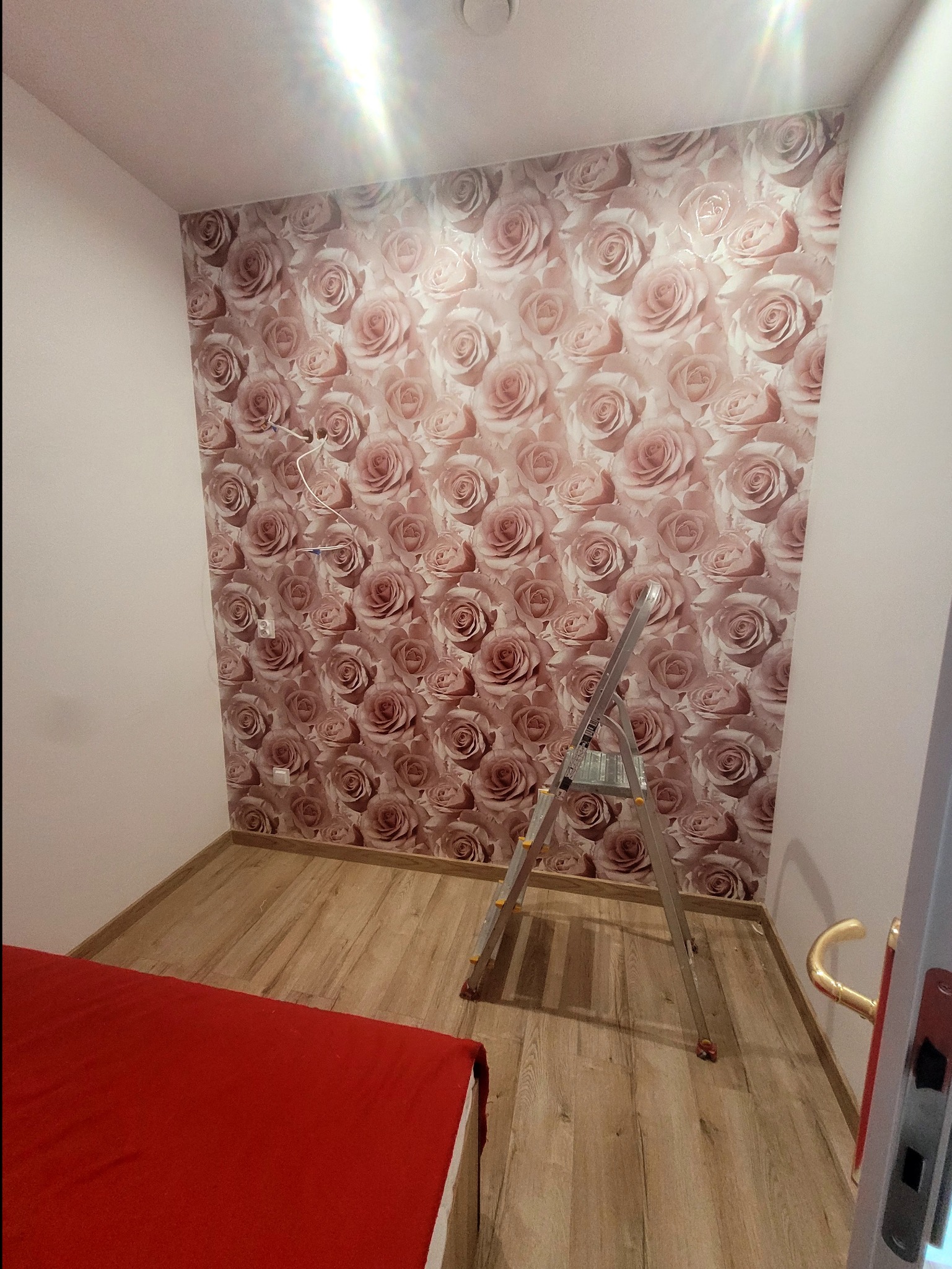 Pokój z tapetą w różowe róże w trakcie remontu, widoczna drabina aluminiowa, gniazdka elektryczne i wystające kable, część czerwonego łóżka w rogu.