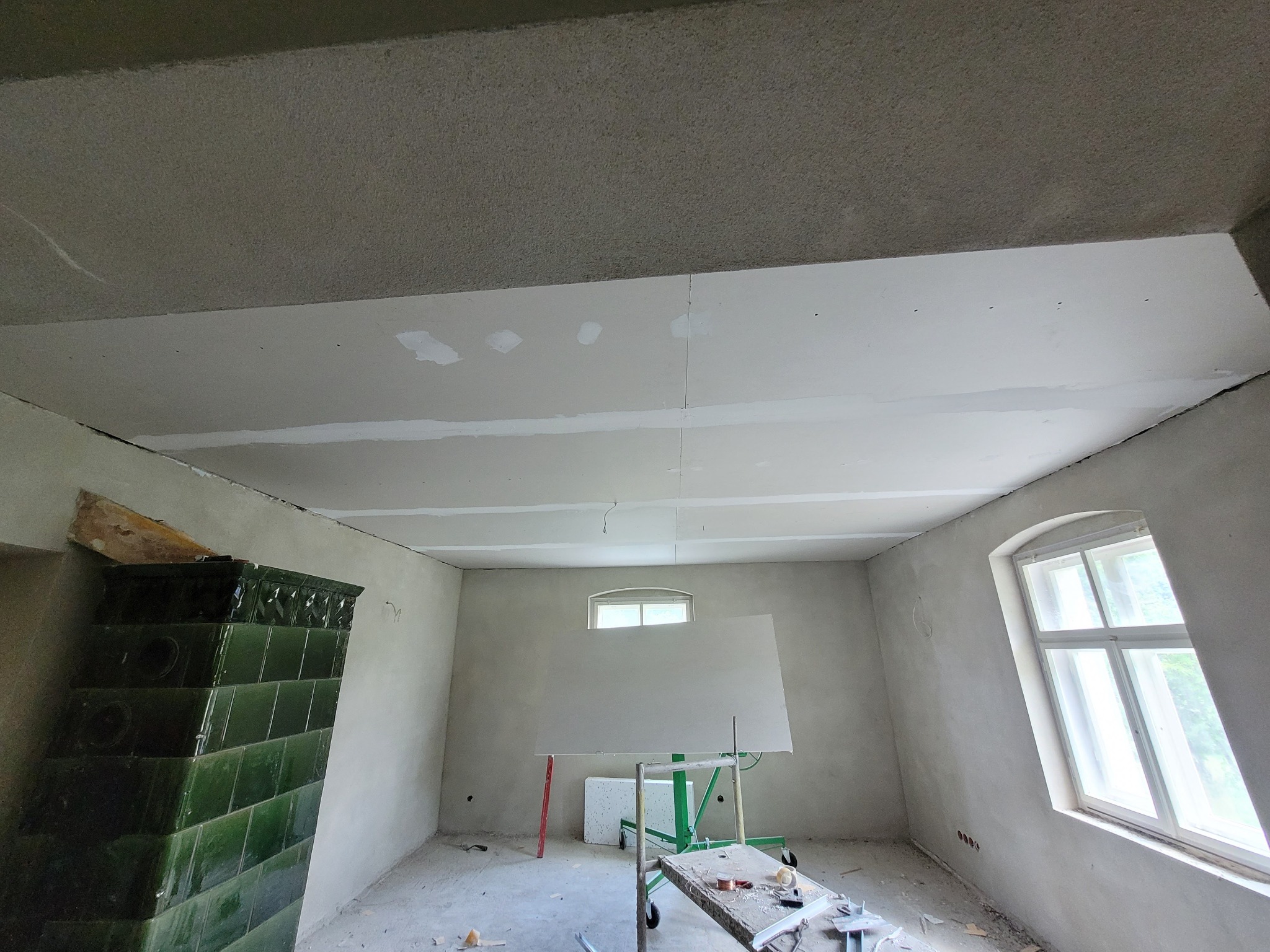 Wnętrze pokoju w trakcie remontu: zamontowana płyta kartonowo-gipsowa na suficie, widoczne łączenia wymagające szpachlowania, stara kaflowa koza w rogu pomieszczenia, okno z łukowym nadprożem...