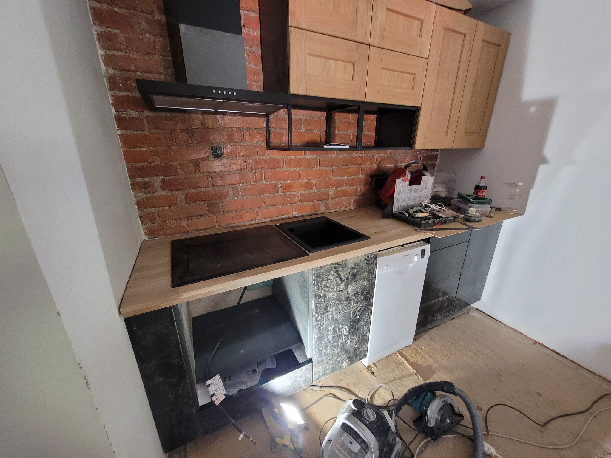 Kuchnia w trakcie remontu: drewniane szafki, czarny okap nad ceglaną ścianą, blat z wbudowanym zlewem i płytą, biała zmywarka, podłoga w trakcie układania, widoczne narzędzia i odkurzacz.