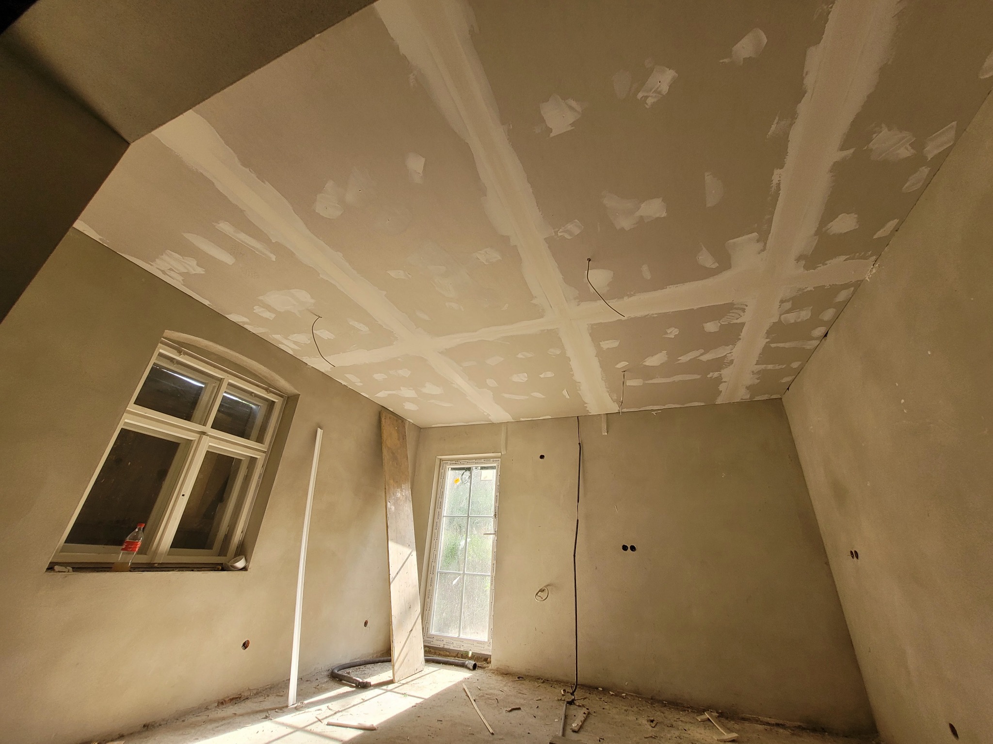 Poddasze w trakcie prac wykończeniowych: widoczne płyty kartonowo-gipsowe na skośnym suficie z nałożoną warstwą szpachli, dwa okna, butelka po napoju w jednym z okien, odsłonięte przewody elektryczne.