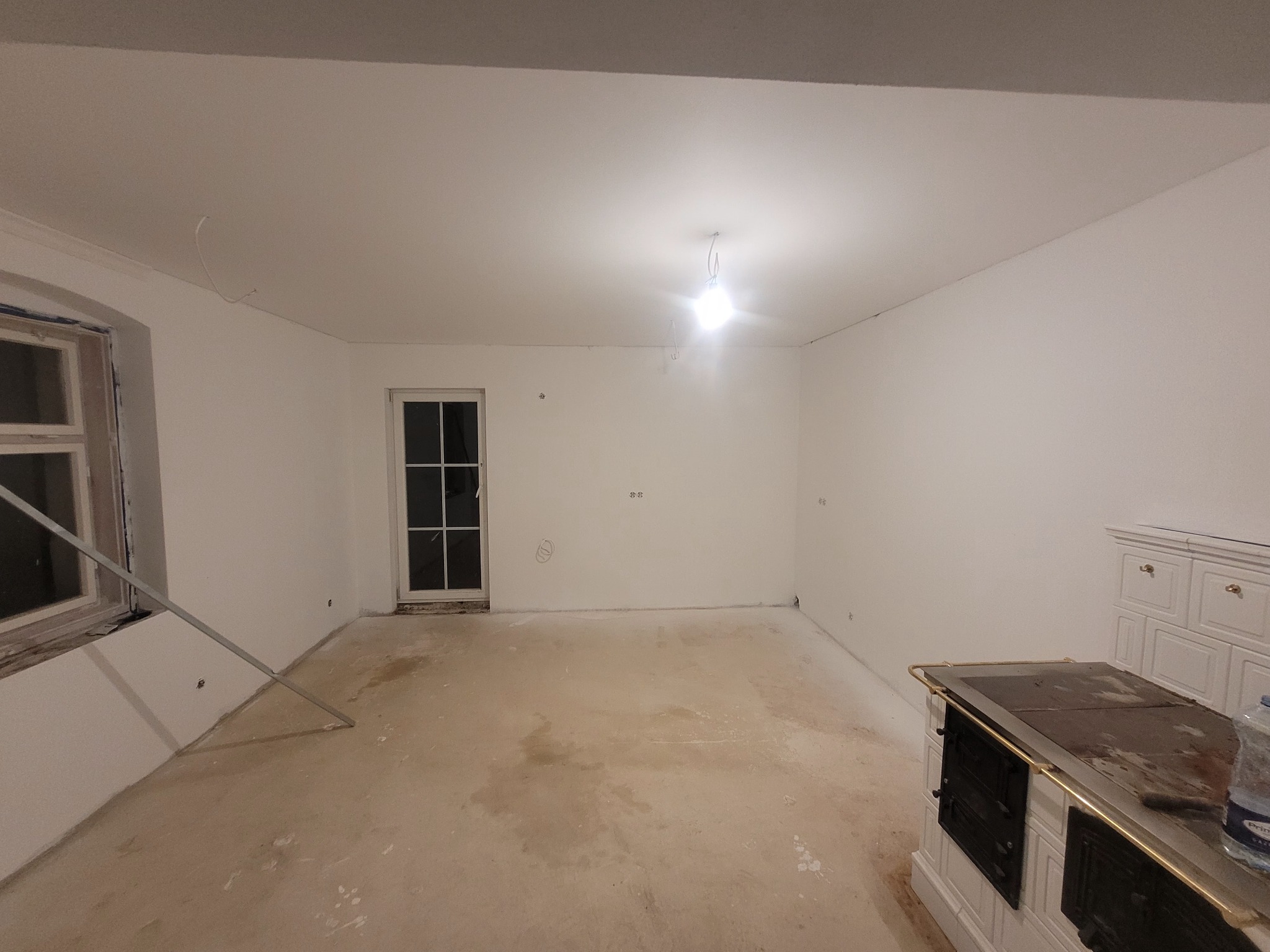 Wnętrze pokoju w trakcie prac wykończeniowych: białe ściany po malowaniu, surowa podłoga przygotowana do dalszych prac, widoczne elementy instalacji elektrycznej oraz piec kaflowy w rogu...