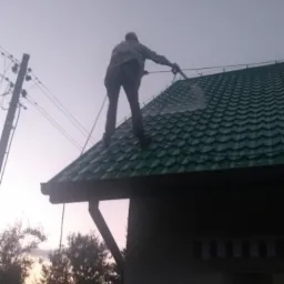 Pracownik na dachu z zielonej blachy trapezowej, trzyma lancę i czyści dach pod lekkim kątem, widoczne przewody elektryczne i drzewa w tle, słabe oświetlenie.