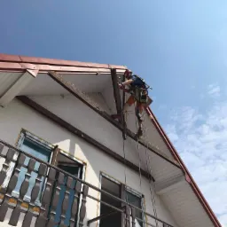 Pracownik w uprzęży, na wysokości, mocuje elementy na dachu budynku dwupiętrowego z balkonem, widok z dołu na błękitne niebo.