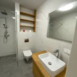 Nowoczesna łazienka z kabiną prysznicową typu walk-in, umywalką nablatową na drewnianej szafce, wiszącą toaletą i drewnianą półką na ścianie wyłożonej białymi płytkami imitującymi marmur.