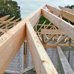 Be Good SP.Z.O.O. - Drewniana konstrukcja dachu w trakcie budowy, widoczne elementy więźby z jasnego drewna na tle muru z bloczków betonowych i zieleni drzew.