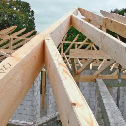Drewniana konstrukcja dachu w trakcie budowy, widoczne elementy więźby z jasnego drewna na tle muru z bloczków betonowych i zieleni drzew.