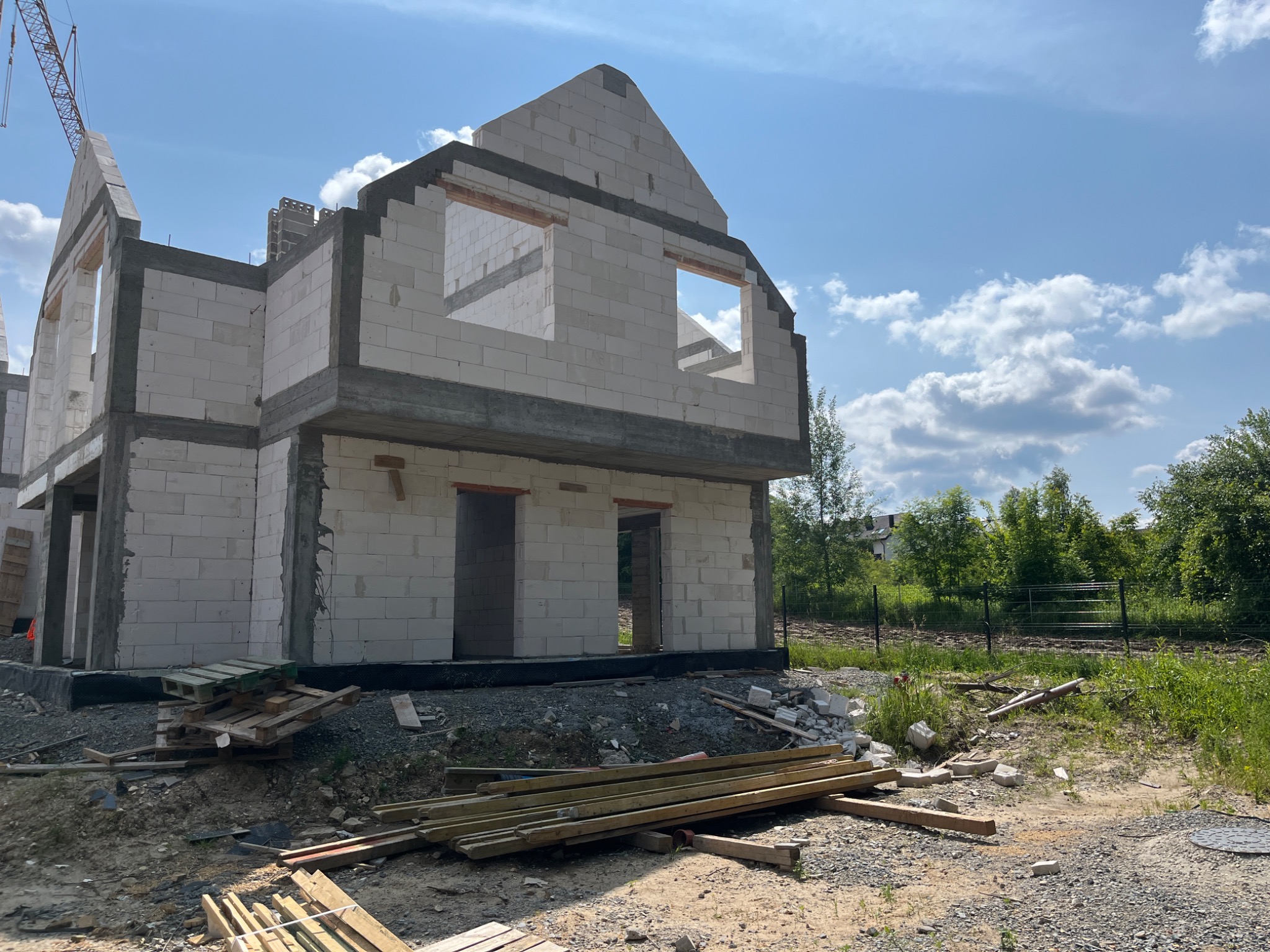 Surowy stan domu z białych bloczków, widoczne otwory okienne i drzwiowe, częściowo zadaszony, na tle błękitnego nieba z chmurami. Elementy budowlane na pierwszym planie.