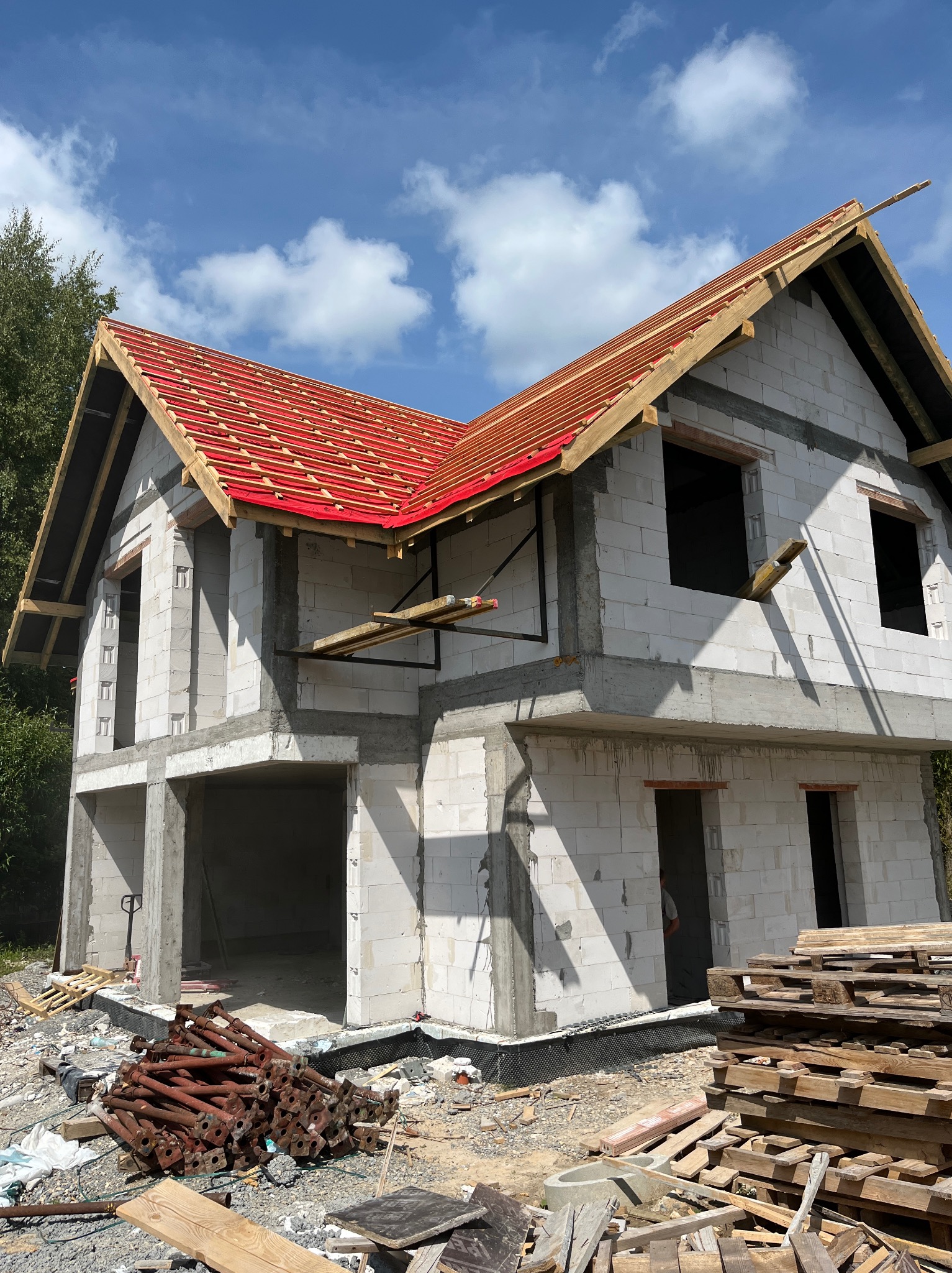 Dom w trakcie budowy z czerwonym dachem, białe bloczki, widoczne rusztowania i palety. Stan surowy otwarty, niebo z chmurami w tle.