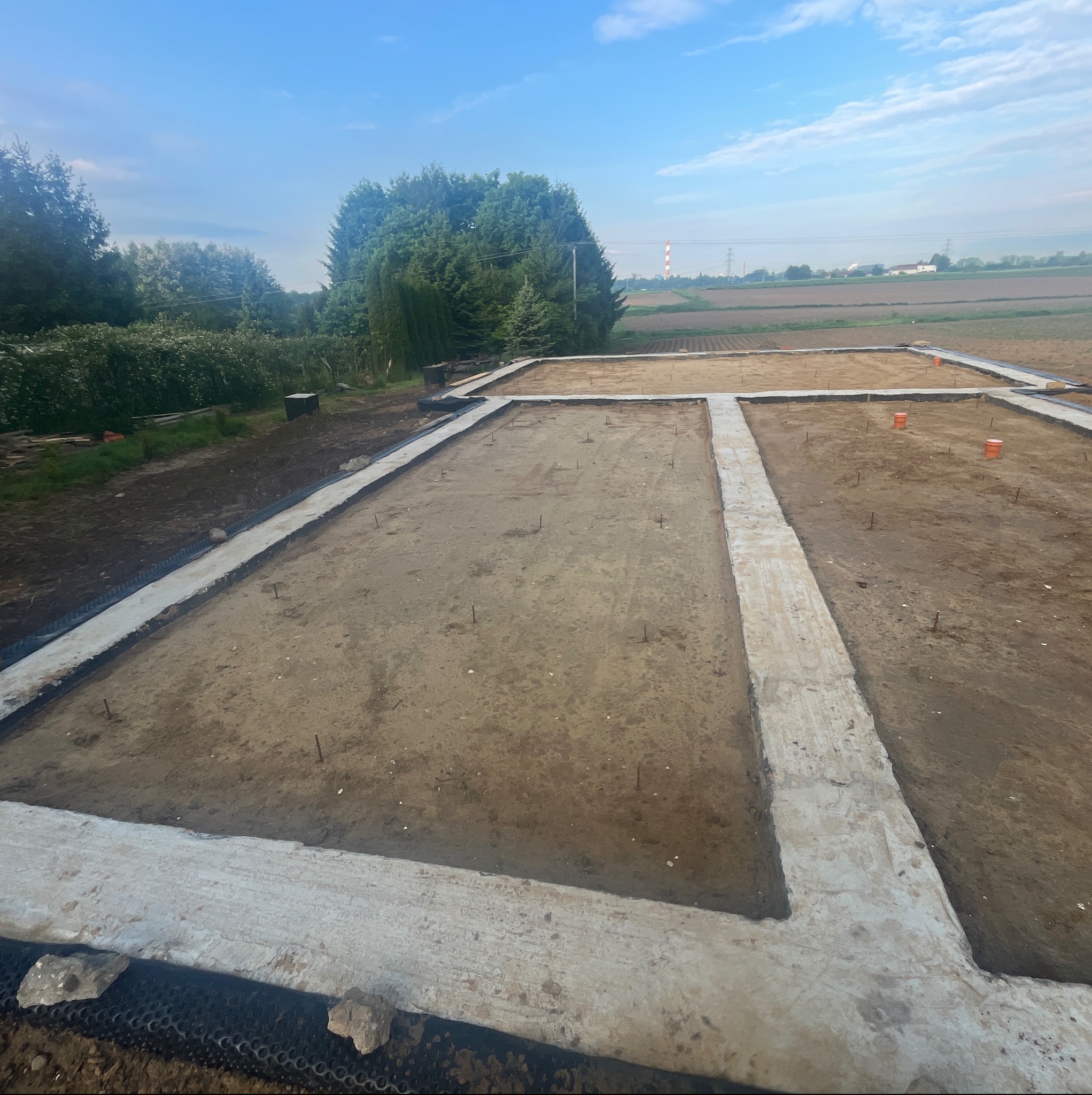 Fundamenty domu w trakcie budowy, widoczne betonowe ławy fundamentowe i teren przygotowany pod wylewkę. W tle zieleń i komin elektrowni.