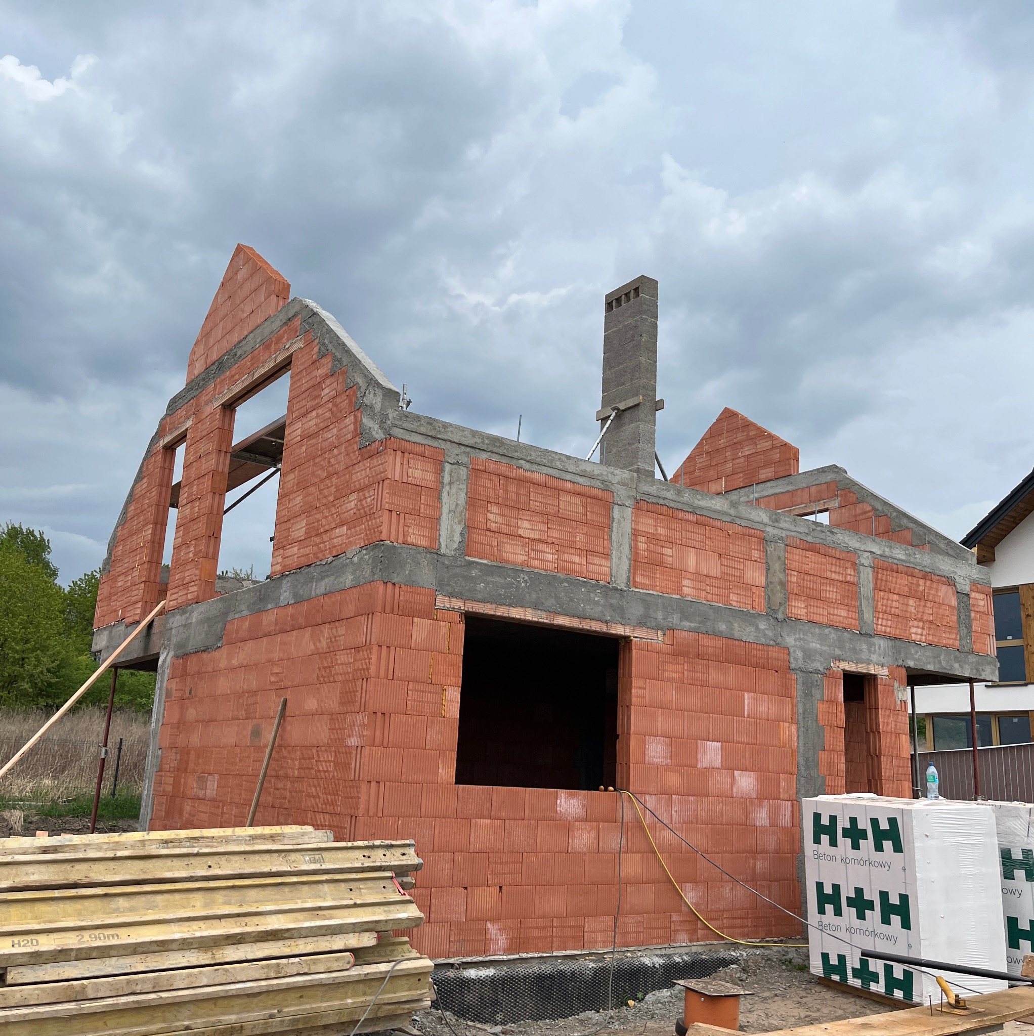 Surowy stan domu z czerwonej cegły H+H, widoczne otwory okienne i komin, w tle pochmurne niebo. Na pierwszym planie leżące deski i paleta z cegłami.