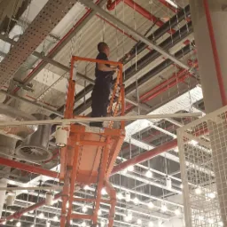 Pracownik na podnośniku koszowym czyści sufit w biurze, widoczne instalacje, rury i oświetlenie.