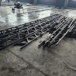 Stalowe kratownice ułożone na betonowej posadzce hali produkcyjnej, widoczny sprzęt spawalniczy w tle.