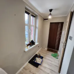 Przedpokój w trakcie malowania ścian na szaro, widoczny kuweta malarska, wałek i pędzel, na parapecie okna leżą różne przedmioty.