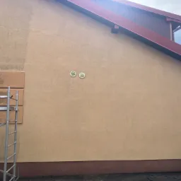 Fragment elewacji budynku w odcieniach beżu, z widoczną różnicą w czystości powierzchni po częściowym umyciu, z dwoma okrągłymi kratkami wentylacyjnymi, fragmentem rusztowania i widocznym...