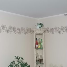 Usługi wykończeniowo-hydrauliczne - Jasny pokój po odświeżeniu: ściany pomalowane na kremowo z dekoracyjnym kwiatowym pasem, biały narożny regał z napojami, firanki w oknie.