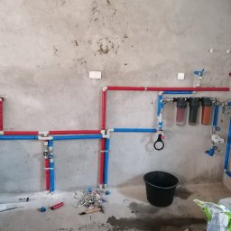 Usługi wykończeniowo-hydrauliczne - Surowa ściana z widoczną instalacją wodną z rur w kolorach czerwonym i niebieskim, filtry wody w przezroczystych obudowach, czarne wiadro i narzędzia na podłodze w trakcie montażu.