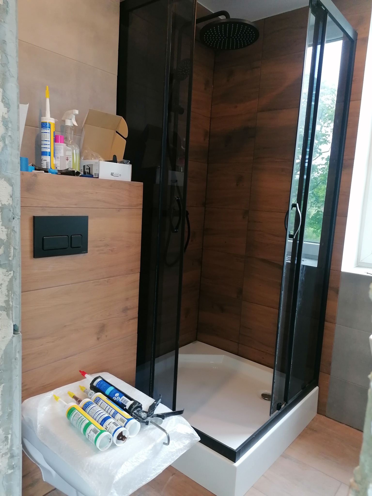 Fragment łazienki w trakcie remontu: kabina prysznicowa z czarnymi ramami, drewnopodobne płytki, zestaw narzędzi i materiałów uszczelniających na sedesie, widoczne niedokończone prace wykończeniowe.