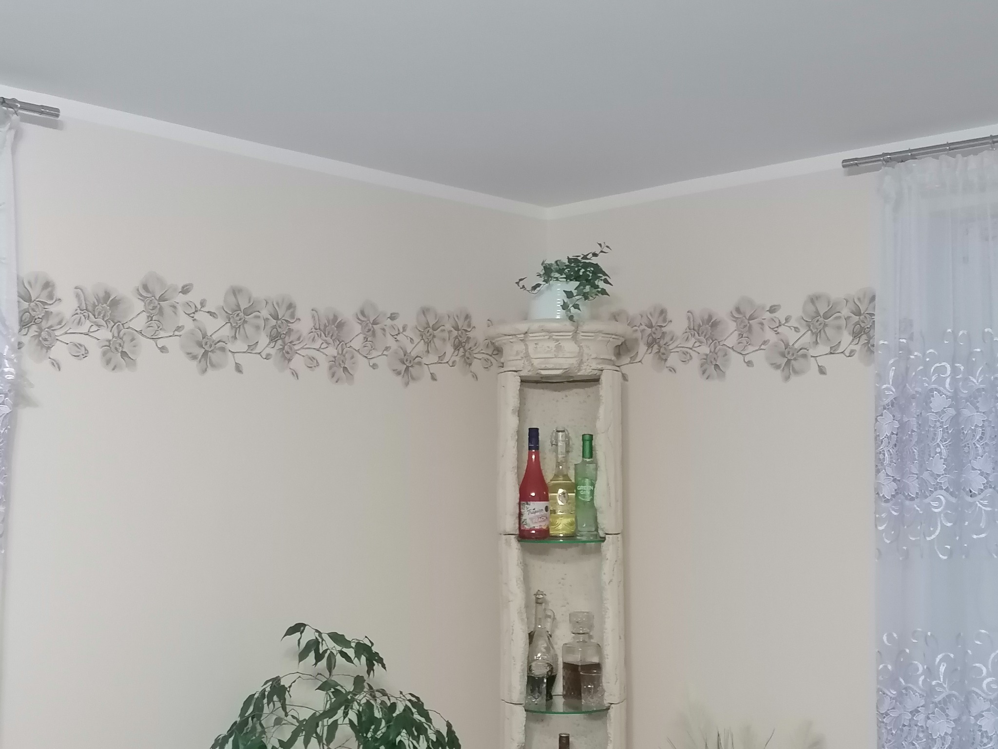 Jasny pokój po odświeżeniu: ściany pomalowane na kremowo z dekoracyjnym kwiatowym pasem, biały narożny regał z napojami, firanki w oknie.