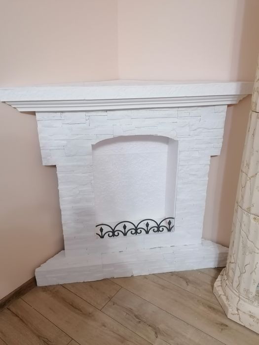 Biały, dekoracyjny kominek z imitacji kamienia, z czarnym, ozdobnym elementem metalowym w palenisku, umieszczony w rogu pokoju na drewnianej podłodze, obok ozdobnej kolumny.