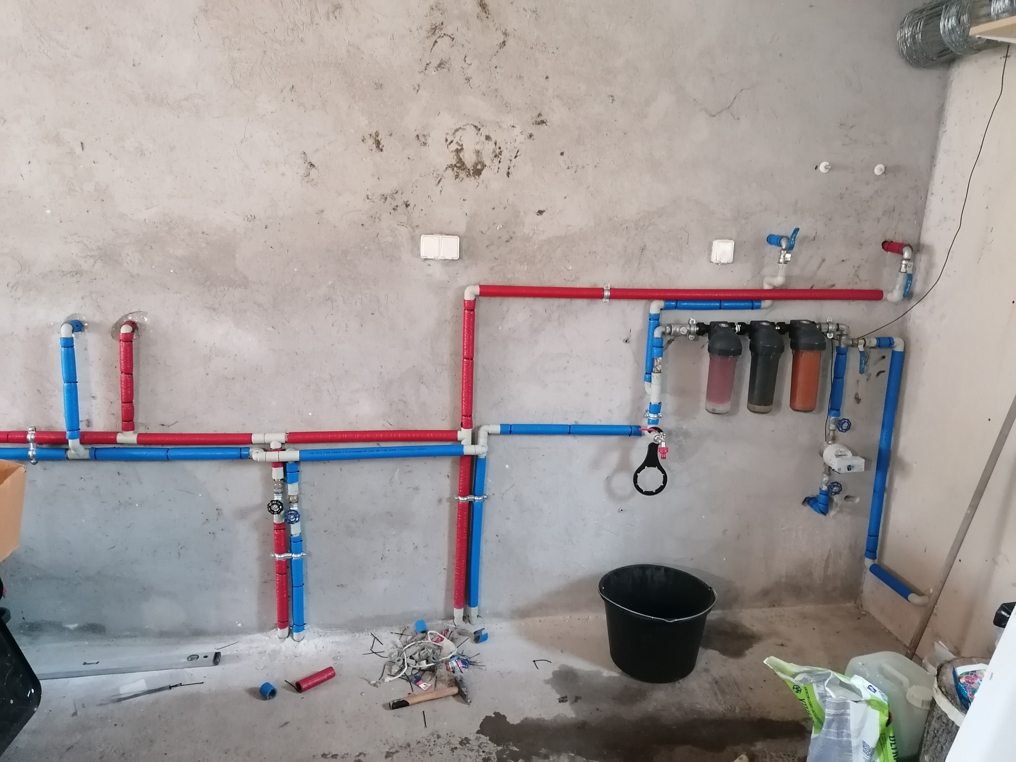 Surowa ściana z widoczną instalacją wodną z rur w kolorach czerwonym i niebieskim, filtry wody w przezroczystych obudowach, czarne wiadro i narzędzia na podłodze w trakcie montażu.