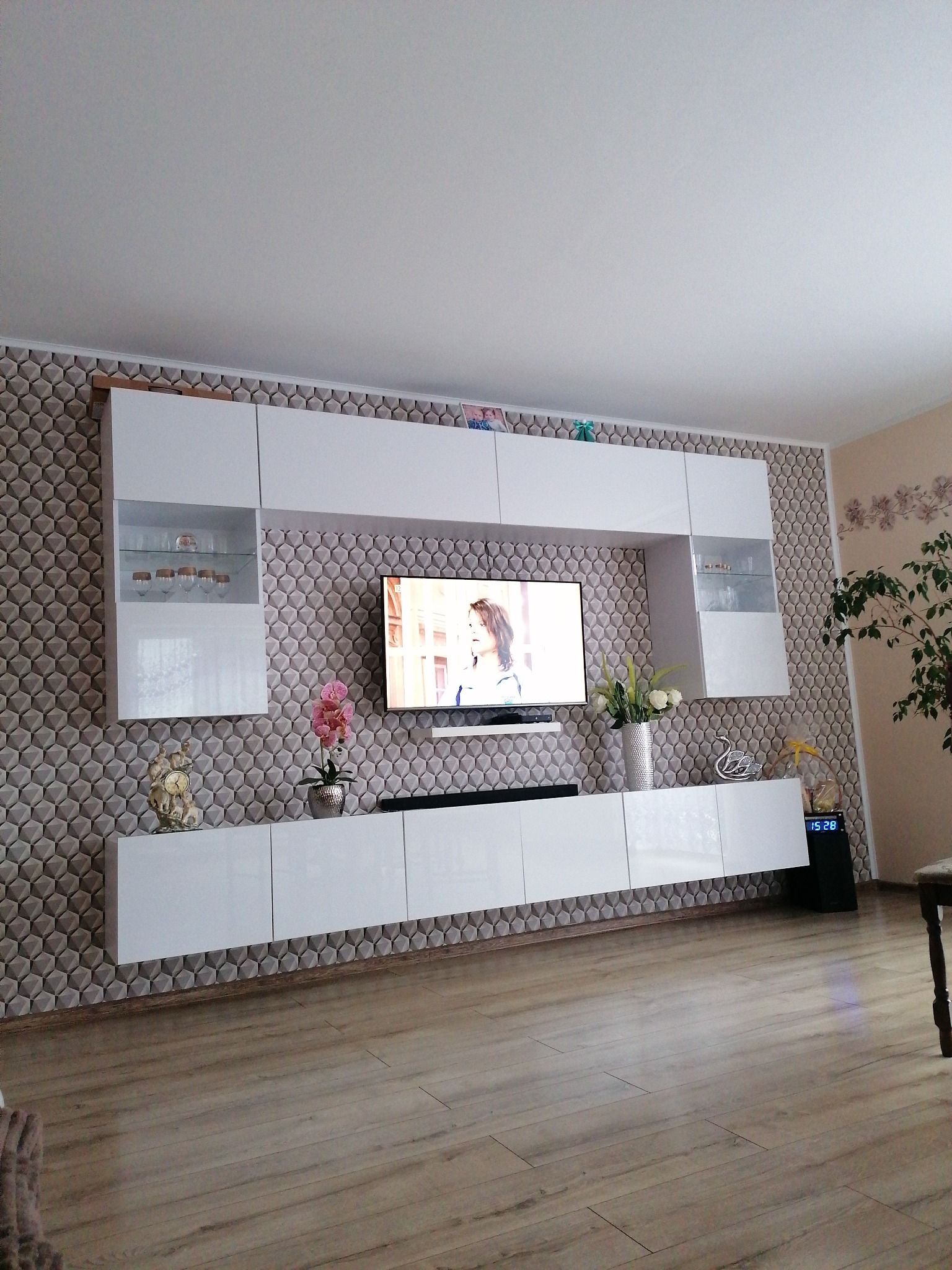 Nowoczesny salon z białą meblościanką na tle ściany z trójwymiarową tapetą, telewizor z widoczną postacią, drewniana podłoga i elementy dekoracyjne.