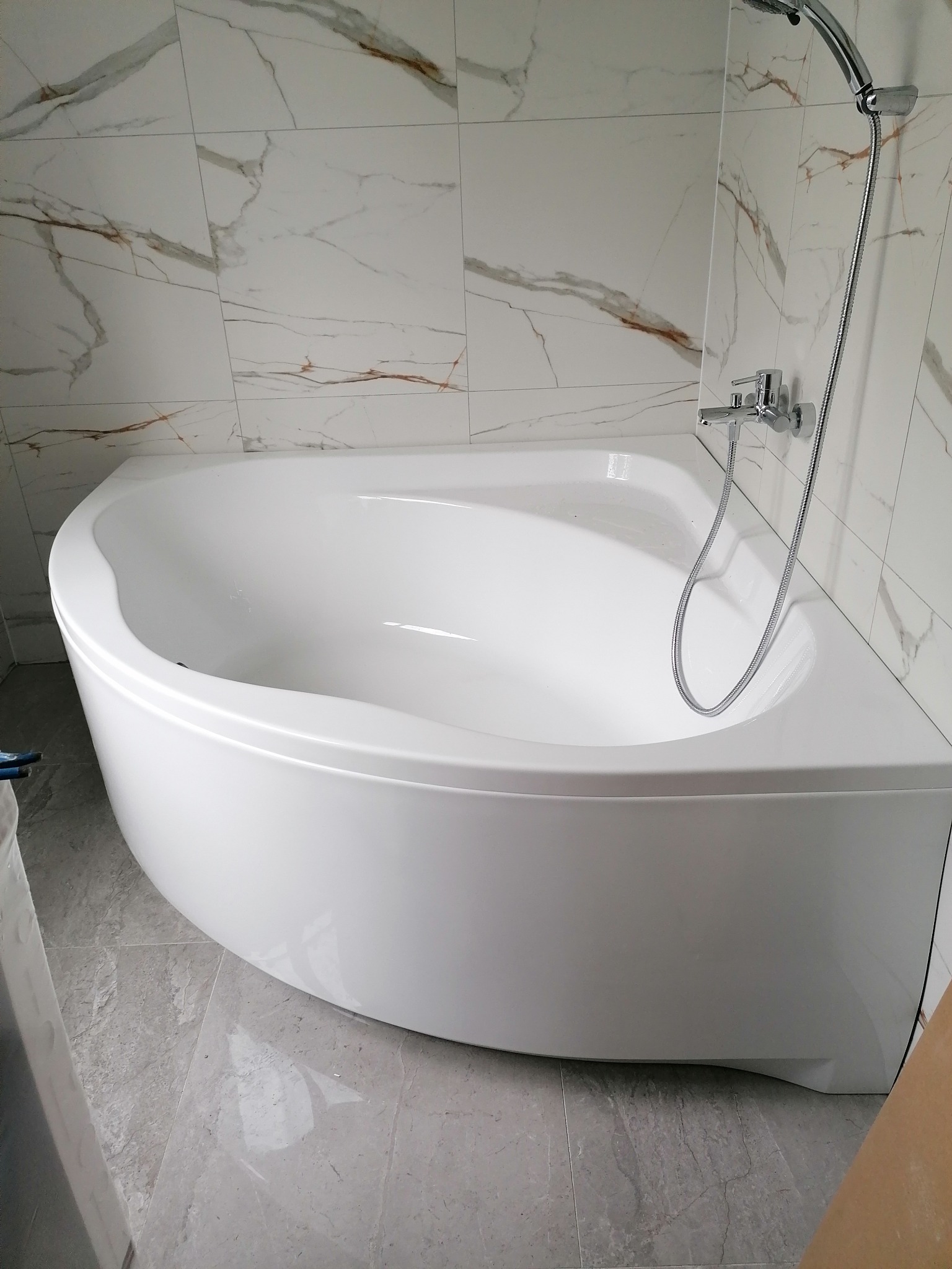 Nowoczesna, narożna wanna z hydromasażem, chromowana bateria i słuchawka prysznicowa na tle ściany z marmurowych płytek, szara podłoga.