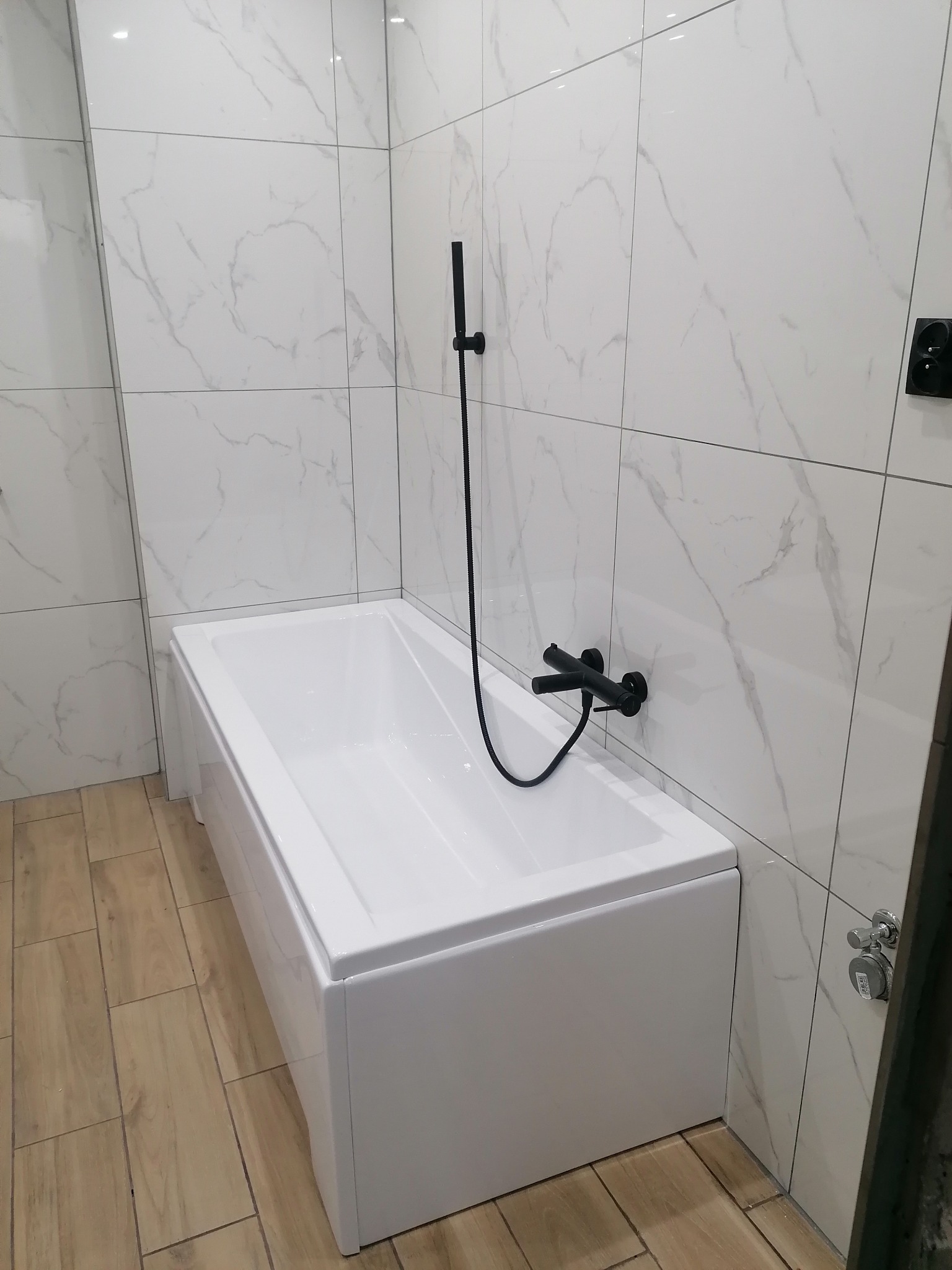 Nowoczesna łazienka z białą wanną, armaturą w kolorze czarnym i płytkami imitującymi marmur na ścianach oraz drewnopodobną podłogą.