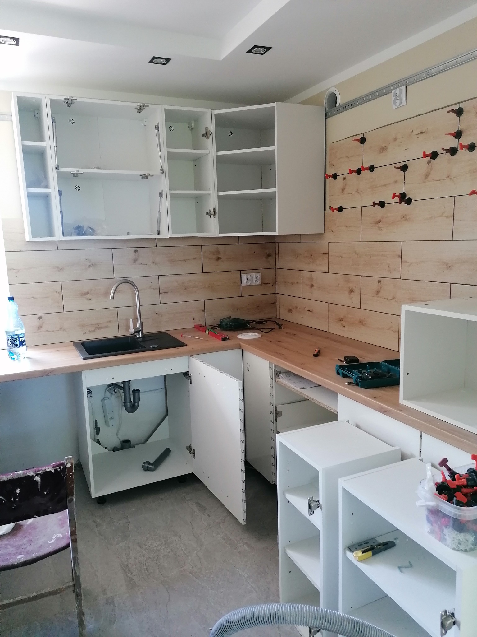 Montaż białej zabudowy kuchennej z drewnopodobnymi płytkami na ścianie, widoczne narzędzia, otwarte szafki i podłączenie hydrauliczne zlewozmywaka.