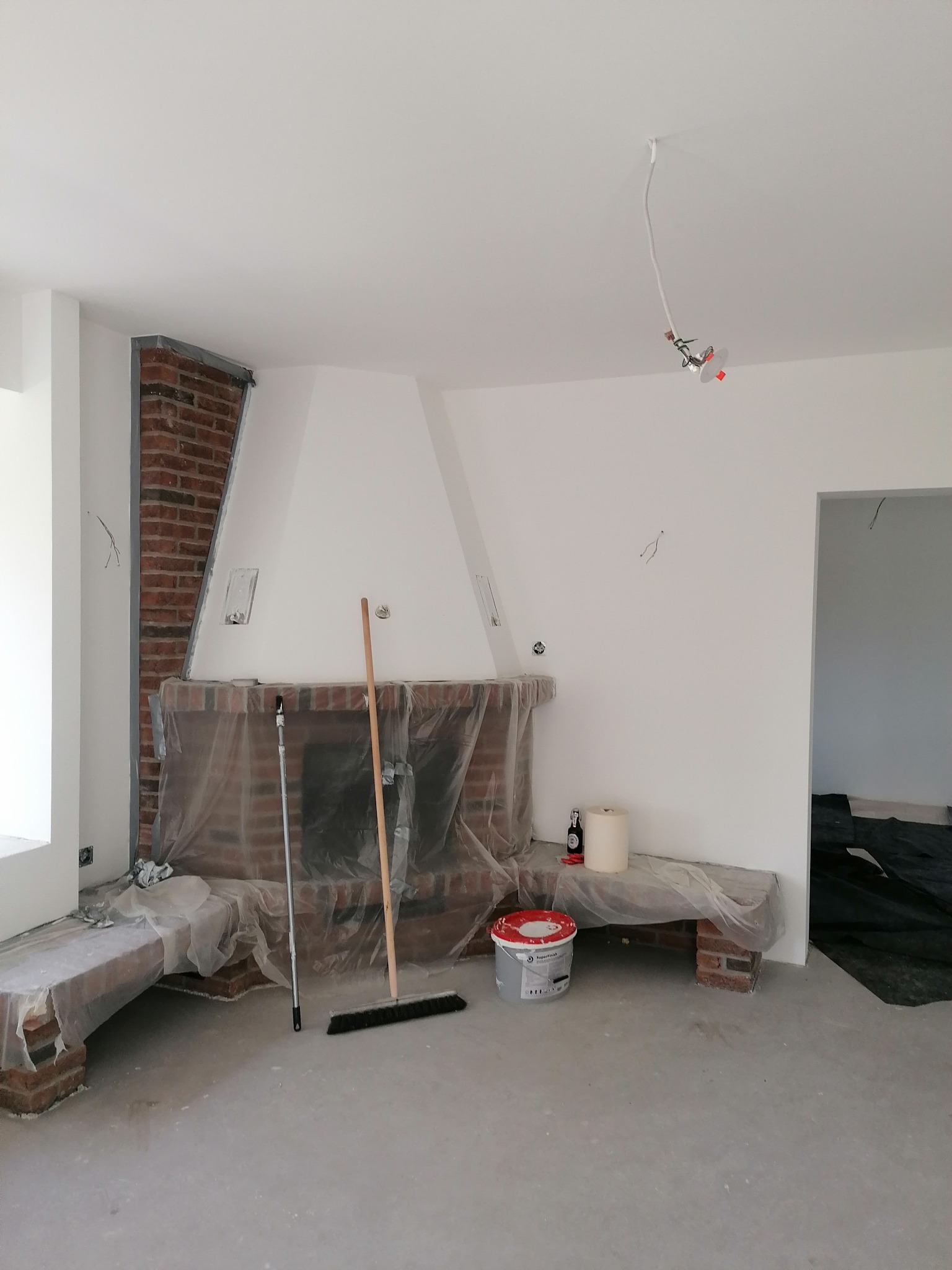 Pokój w trakcie malowania: zabezpieczony kominek z cegły, białe ściany, wiadro z farbą SuperFrank na podłodze, widoczne przewody elektryczne wystające z sufitu.