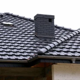 Czarny dach z dachówki ceramicznej z kominem z cegły klinkierowej i oknem dachowym, widoczne rynny i fragment ściany budynku w trakcie budowy.