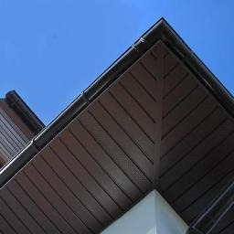 Fragment okapu dachu z brązową podbitką i rynną w Koninie, sfotografowany pod kątem na tle błękitnego nieba.