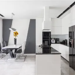 Nowoczesna kuchnia z białymi szafkami, czarnym blatem, wyspą kuchenną i stołem jadalnym z bukietem słonecznik&oacute;w w czarnym wazonie.