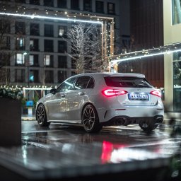 Julian Dzwonkowski - Srebrny samochód marki Mercedes-Benz klasy A, widziany od tyłu na mokrym asfalcie w nocy, z widocznymi światłami tylnymi i tablicą rejestracyjną, na tle oświetlonego budynku.
