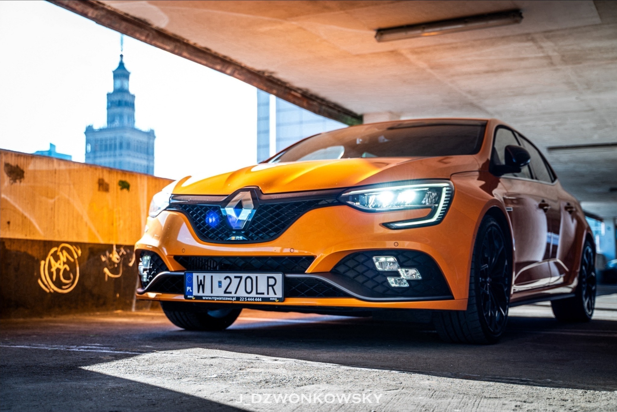 Pomarańczowy samochód Renault Megane RS zaparkowany w garażu podziemnym, z Pałacem Kultury w tle.