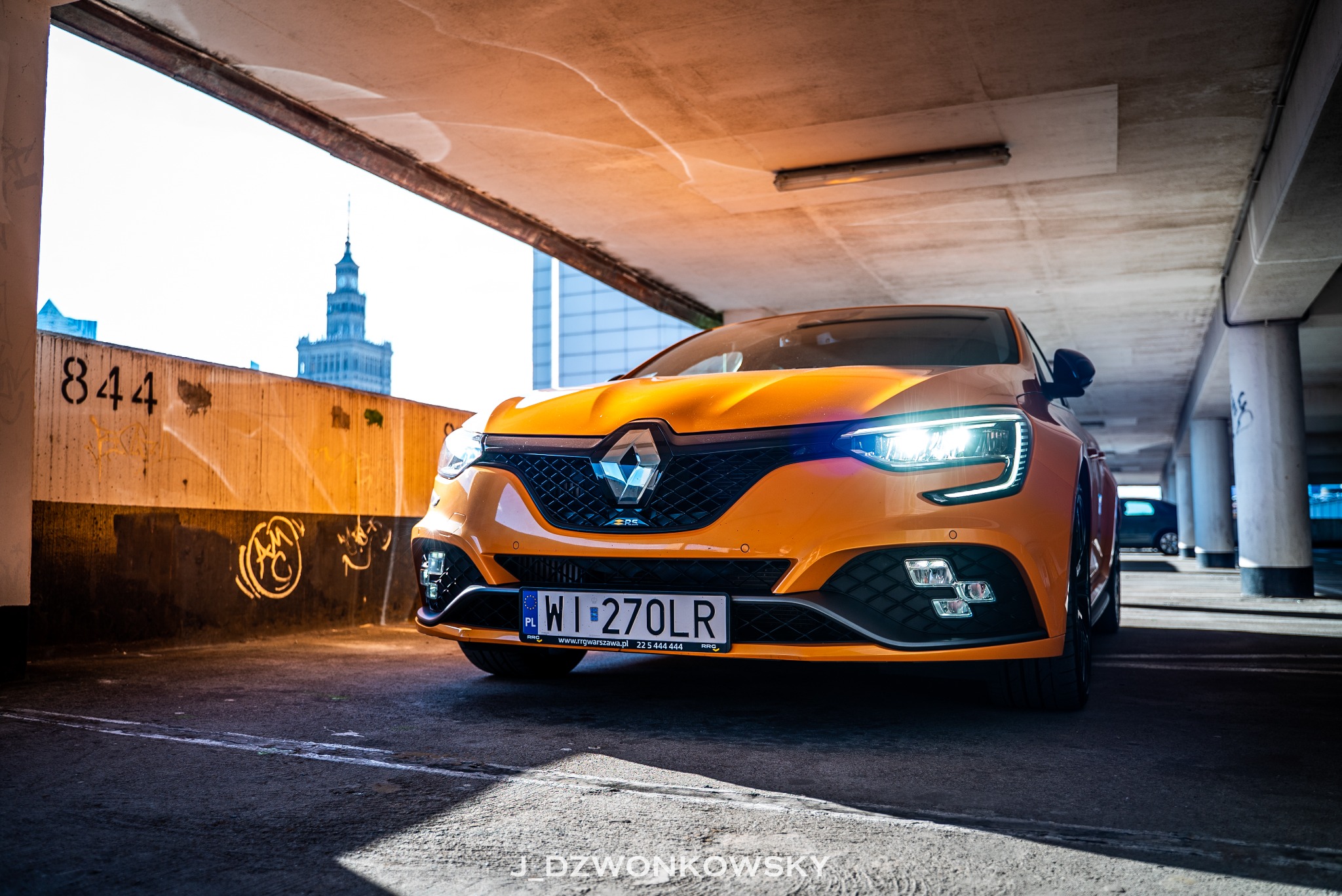 Pomarańczowy Renault Megane RS zaparkowany w garażu podziemnym, z widokiem na Pałac Kultury w tle.